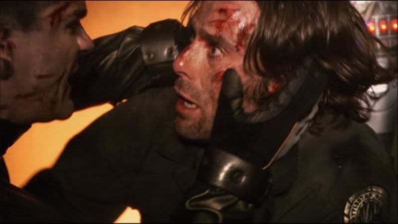 Picture of a scene from a Battlestar Galactica episode. A guy, Gaius Baltar, has just emerged from a burning plane and looks completely lost in thought. Another guy has grabbed Gaius's face and is yelling at him to try to get his attention.