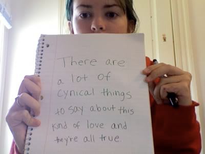 a picture of me, a 21-year-old woman wearing a t-shirt and sweatshirt, holding up a notebook opened to a page that says: There are a lot of cynical things to say about this kind of love and they're all true